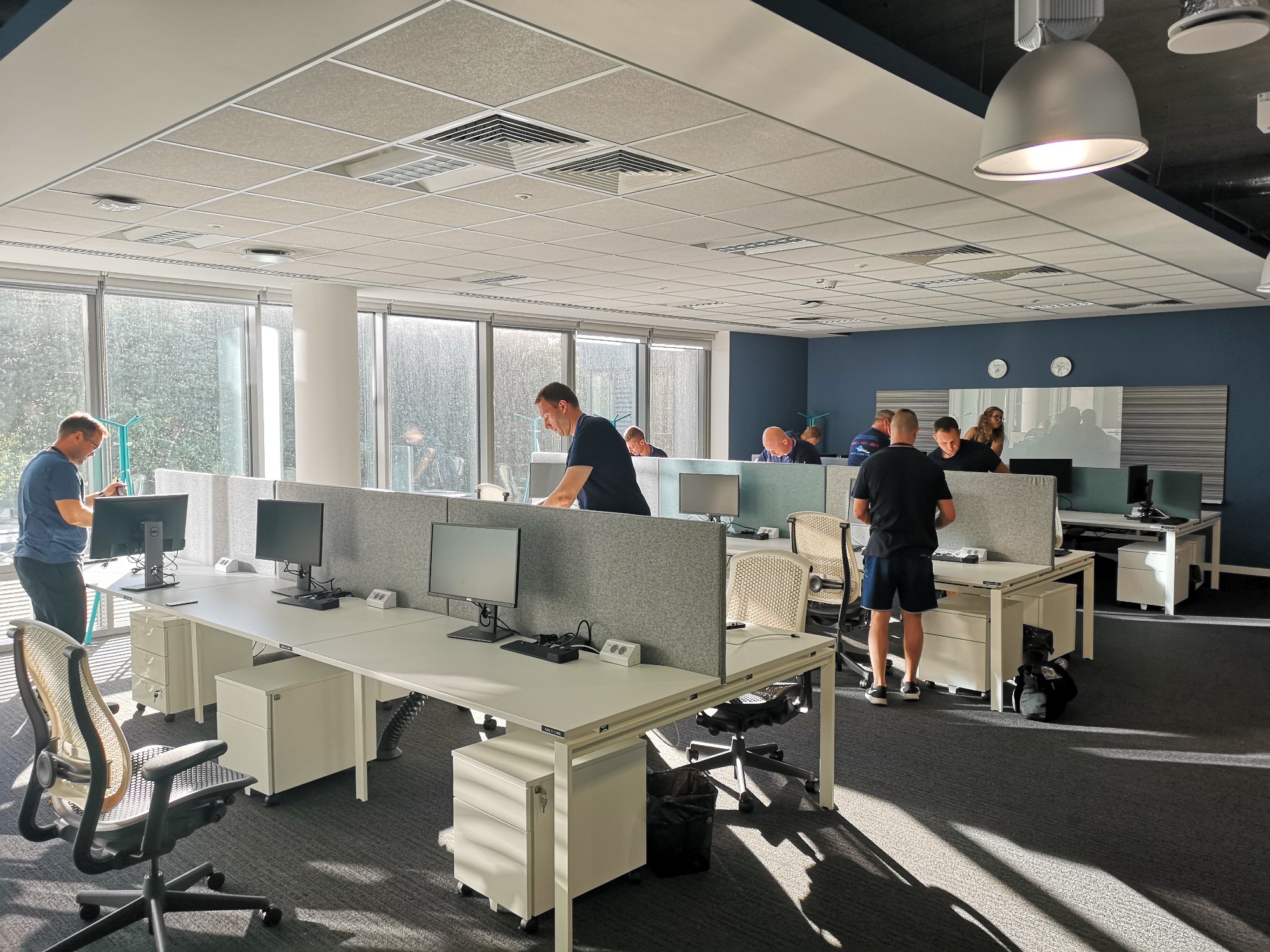 Przestronne biuro typu open space z jasnymi stanowiskami pracy, pracownikami przy komputerach oraz dużymi oknami zapewniającymi naturalne światło.