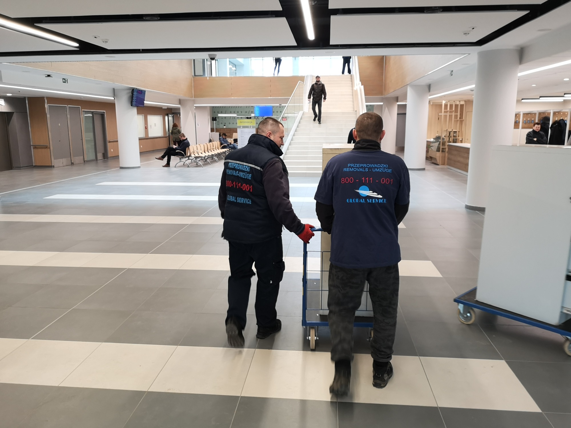 Dwóch pracowników firmy przeprowadzkowej transportuje kartony na wózku w przestronnym, nowoczesnym budynku z białymi kolumnami i schodami.