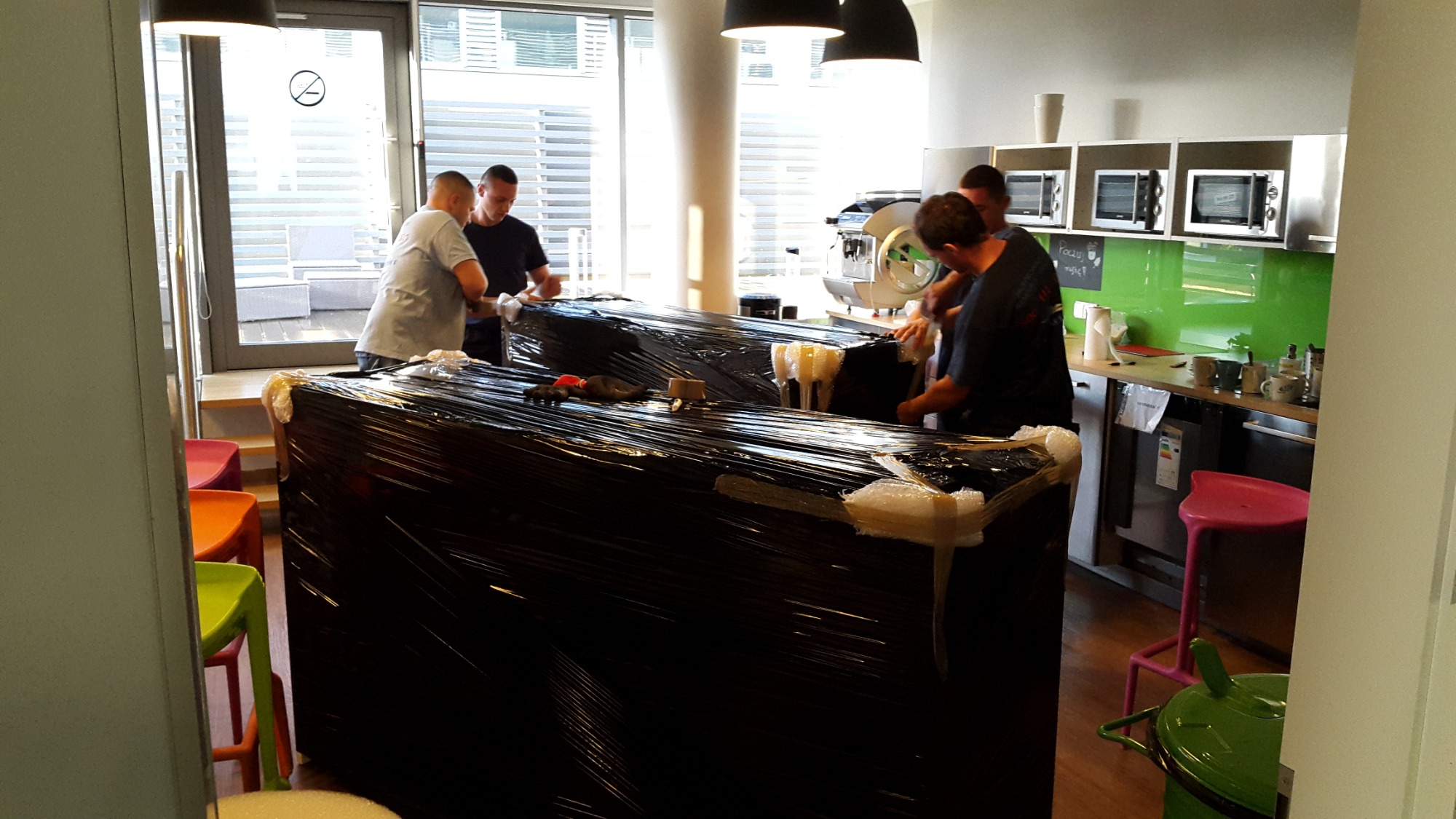 Czterech mężczyzn owija folią stretch meble biurowe przed przeniesieniem w jasnym pomieszczeniu biurowym z aneksem kuchennym.