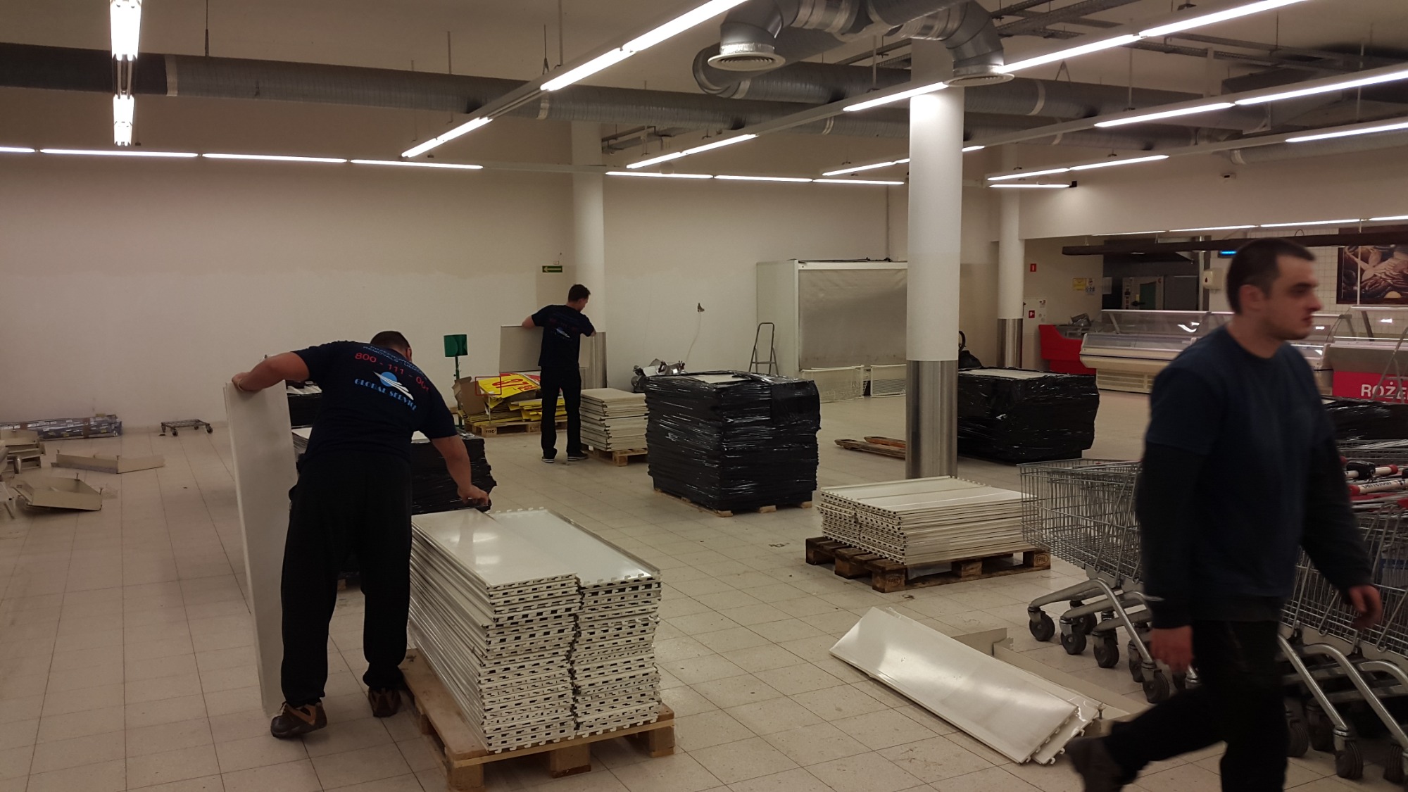 Ekipa demontuje lub montuje wyposażenie sklepu, widoczne stosy białych półek na paletach, w tle mężczyzna w niebieskiej koszulce z logo firmy transportowej