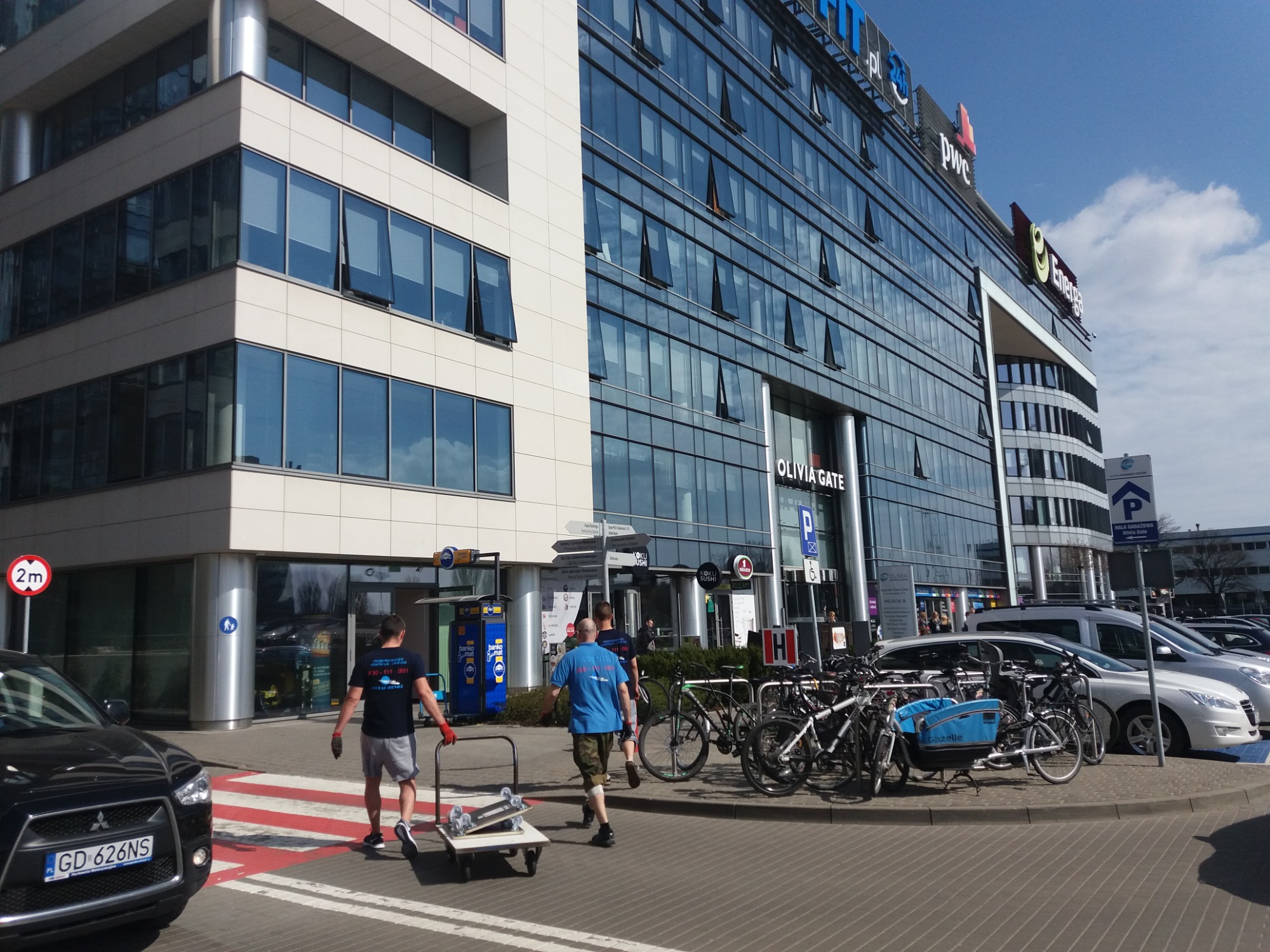 Dwóch mężczyzn transportuje wózek z deskorolką na tle nowoczesnego biurowca Olivia Gate w słoneczny dzień.
