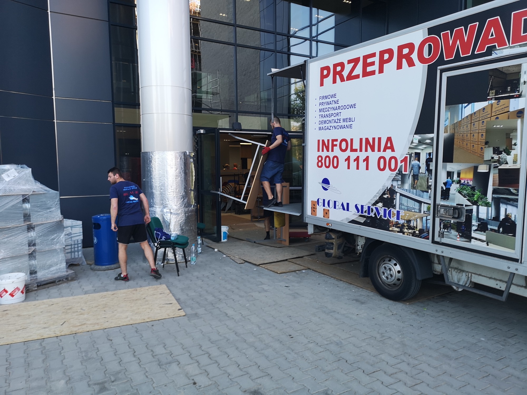 Pracownicy firmy przeprowadzkowej w trakcie załadunku mebli do samochodu na tle nowoczesnego budynku biurowego; widoczny fragment oklejonego samochodu z napisem 'Przeprowadzki' i numerem infolinii.