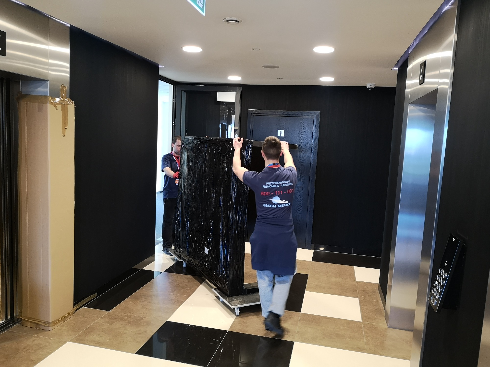 Dwóch pracowników w niebieskich koszulkach z logo firmy transportowej przewozi na wózku owinięty czarną folią mebel w nowoczesnym budynku z windami i czarno-białą podłogą.