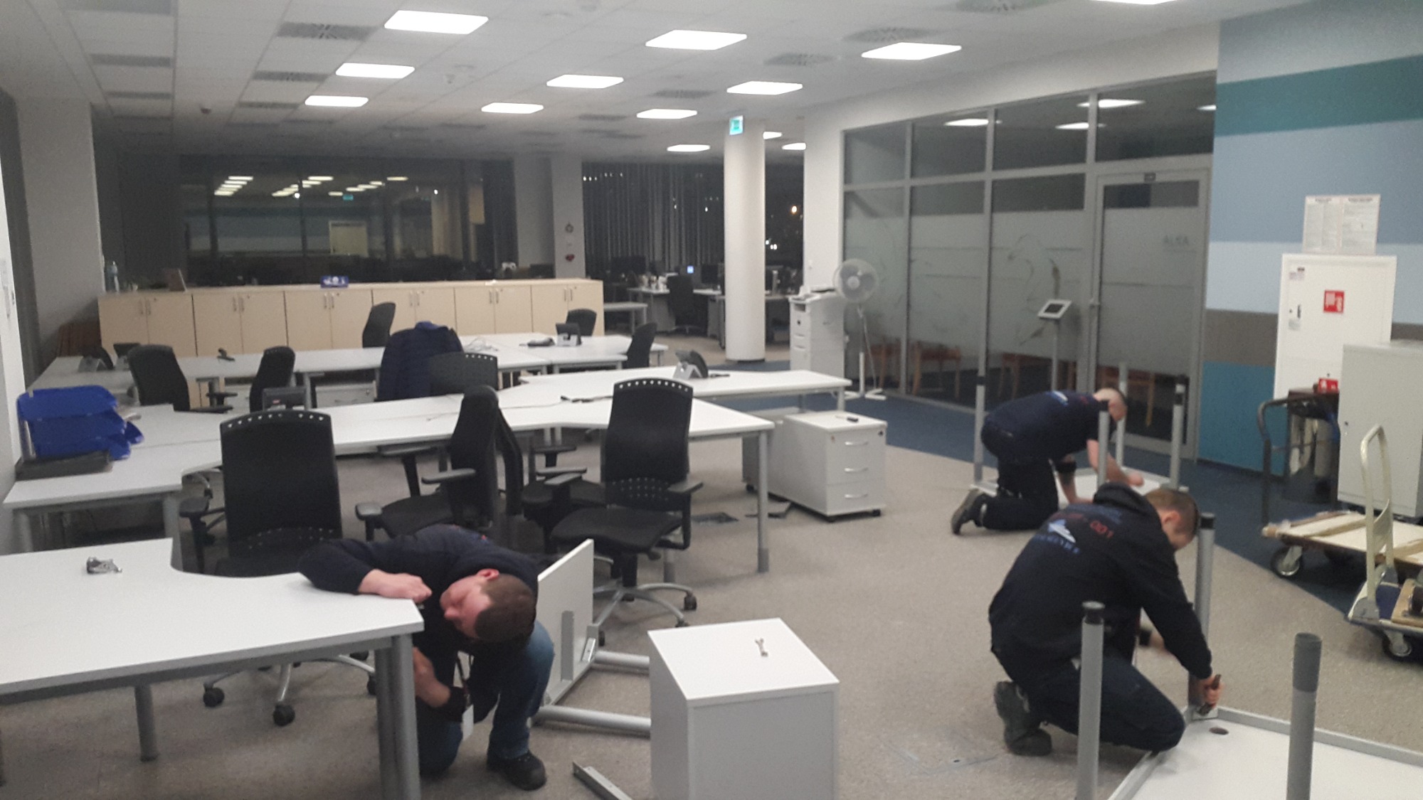 Montaż mebli biurowych w przestronnym pomieszczeniu z widokiem na nocne miasto, dwóch monterów w ciemnych bluzach z logo firmy skręca metalowe nogi biurka, trzeci monter pochyla się nad blatem...