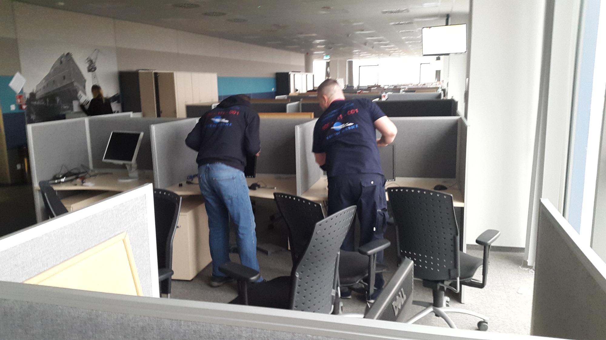 Dwóch mężczyzn w granatowych bluzach z logo firmy demontuje stanowiska pracy w biurze z szarymi boksami i komputerami.
