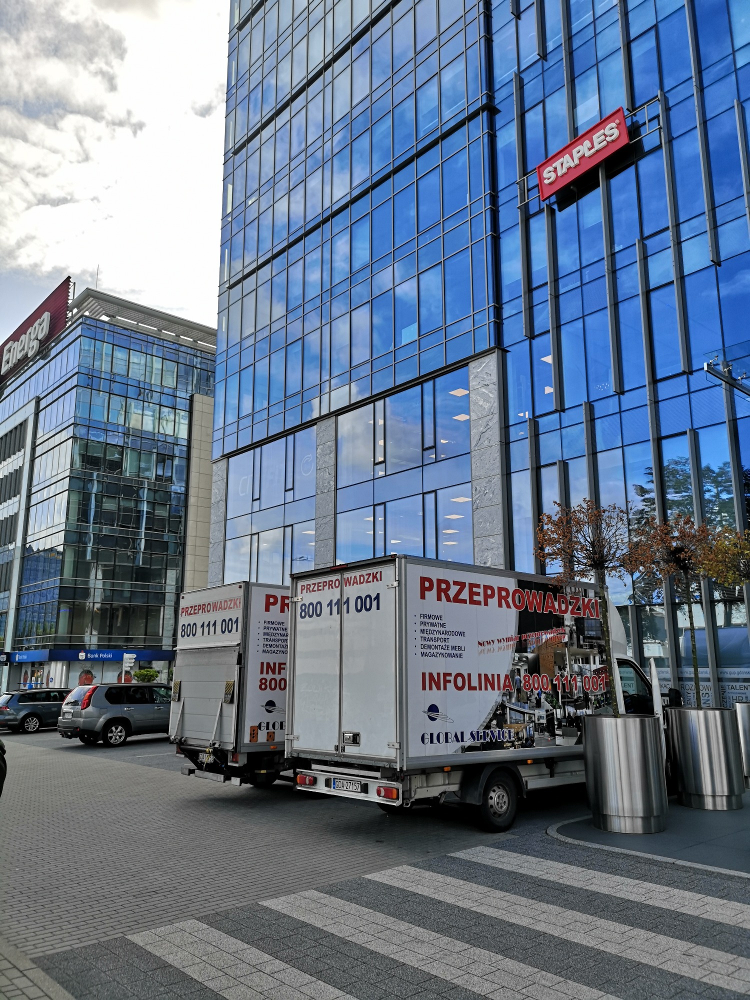 Dwa białe busy z napisem PRZEPROWADZKI zaparkowane przed nowoczesnym biurowcem z logo Staples i Energa, widoczne z poziomu przejścia dla pieszych.