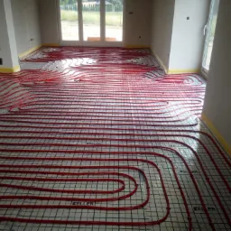 Czerwone rury ogrzewania podłogowego ułożone na czarnej siatce w surowym wnętrzu, żółta taśma zabezpieczająca przy ścianach, widoczne okna.