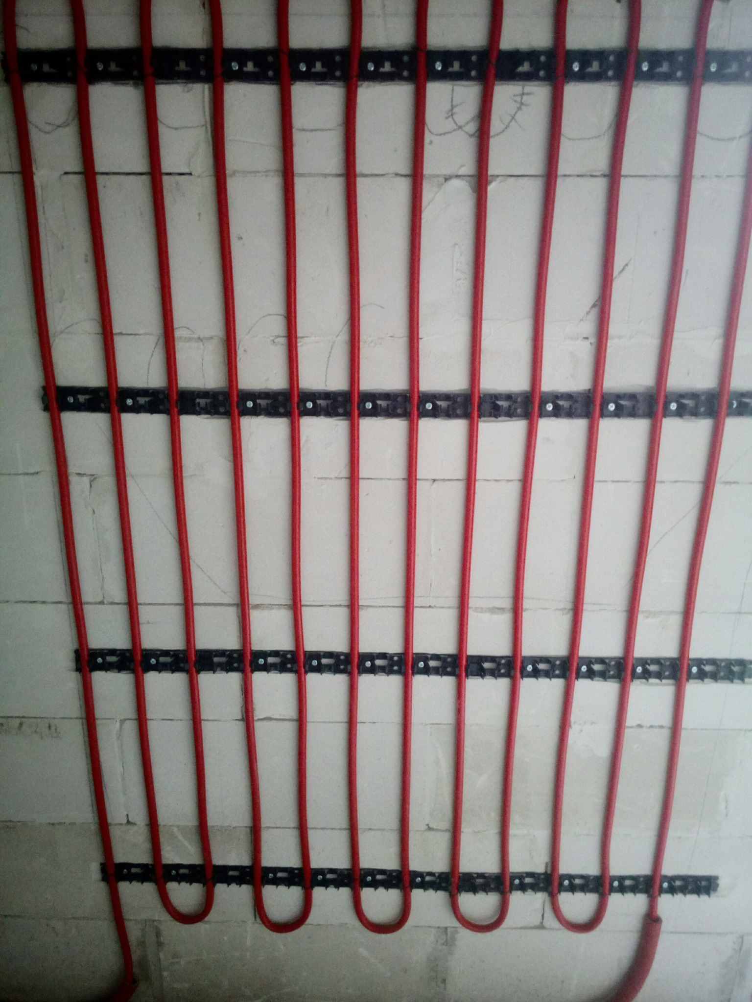 Czerwone rury ogrzewania podłogowego zamocowane na ścianie z białych bloczków, widoczne czarne listwy montażowe z metalowymi klipsami.