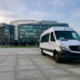 Biały bus Mercedes-Benz Sprinter na tle nowoczesnego biurowca z polską flagą, na placu wyłożonym szarą kostką.