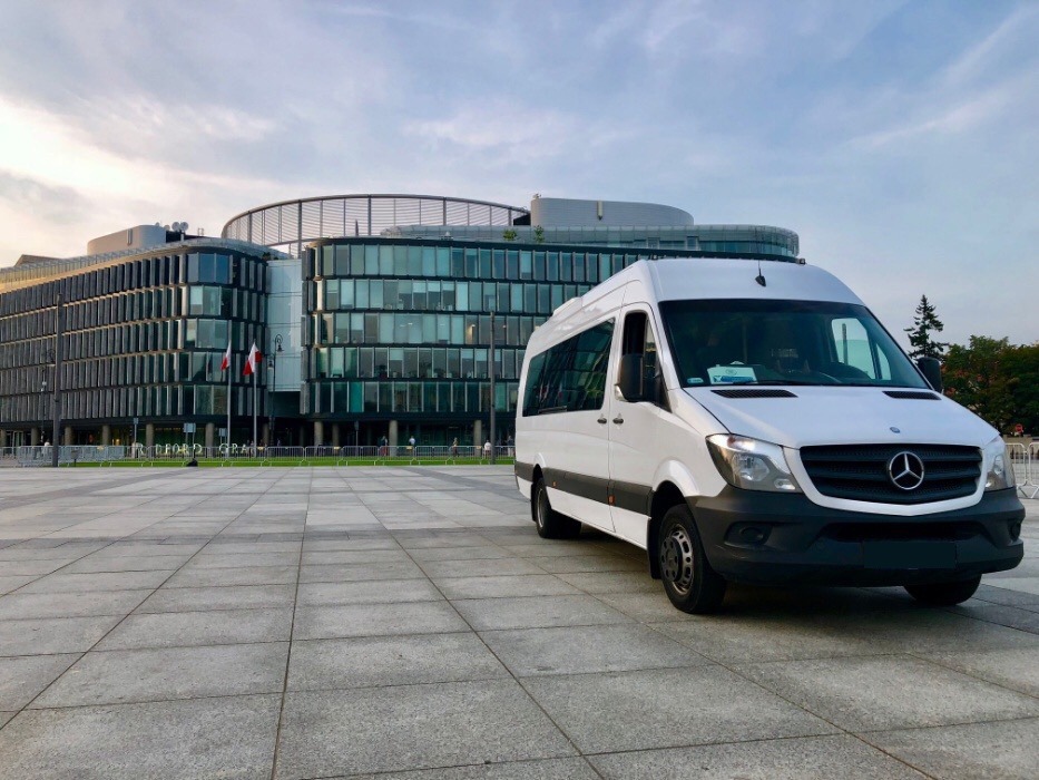 Biały bus Mercedes-Benz Sprinter na tle nowoczesnego biurowca z polską flagą, na placu wyłożonym szarą kostką.