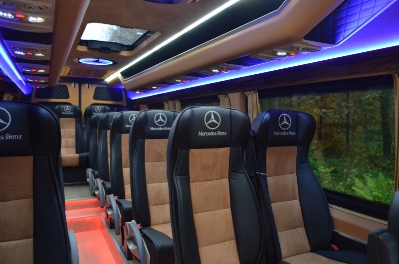 Wnętrze luksusowego busa Mercedes-Benz z czarno-brązowymi skórzanymi fotelami, podświetleniem LED i widokiem na las za oknem.