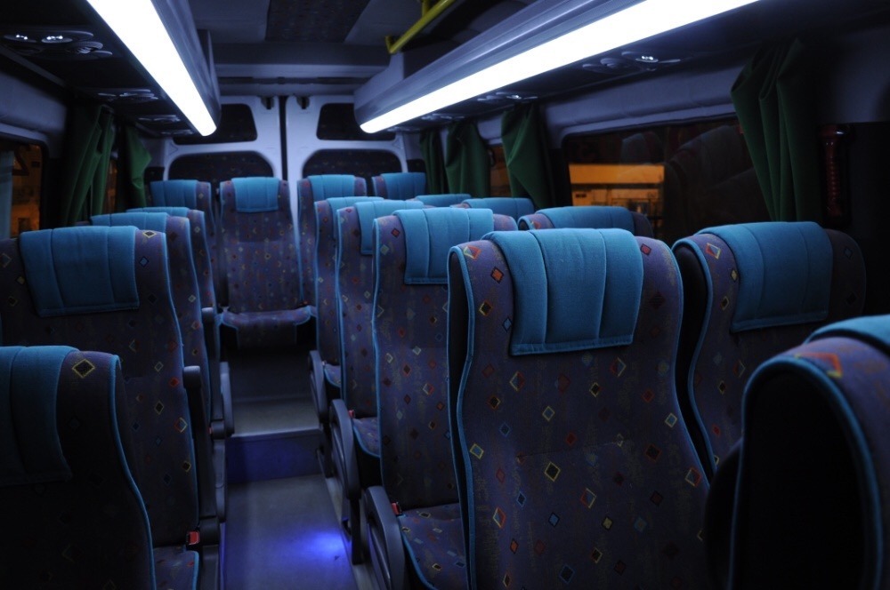 Wnętrze autobusu z rzędami niebiesko-wzorzystych siedzeń podłużnie oświetlonych lampami LED.