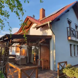 Jasnoszary dom jednorodzinny z czerwoną dachówką, drewnianą werandą i balkonem, otoczony zielonym ogrodem w słoneczny dzień.