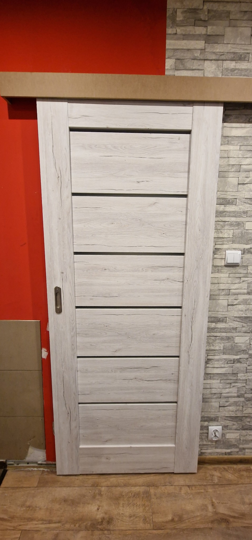 Białe, przesuwne drzwi z poziomymi panelami, zamontowane między czerwoną ścianą a ścianą z imitacji kamienia, widoczny fragment podłogi i gniazdko elektryczne.