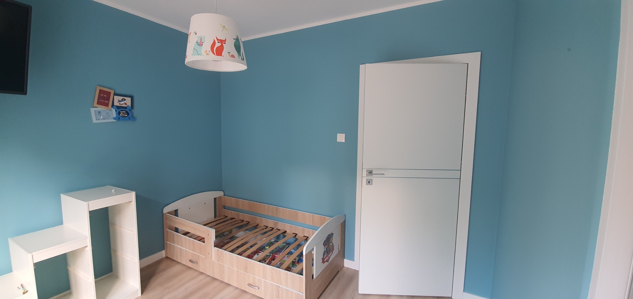 Pomieszczenie dziecięce z błękitnymi ścianami, łóżeczkiem z motywem bajkowym, białą szafką i drzwiami, oświetlone lampą z rysunkami zwierząt.