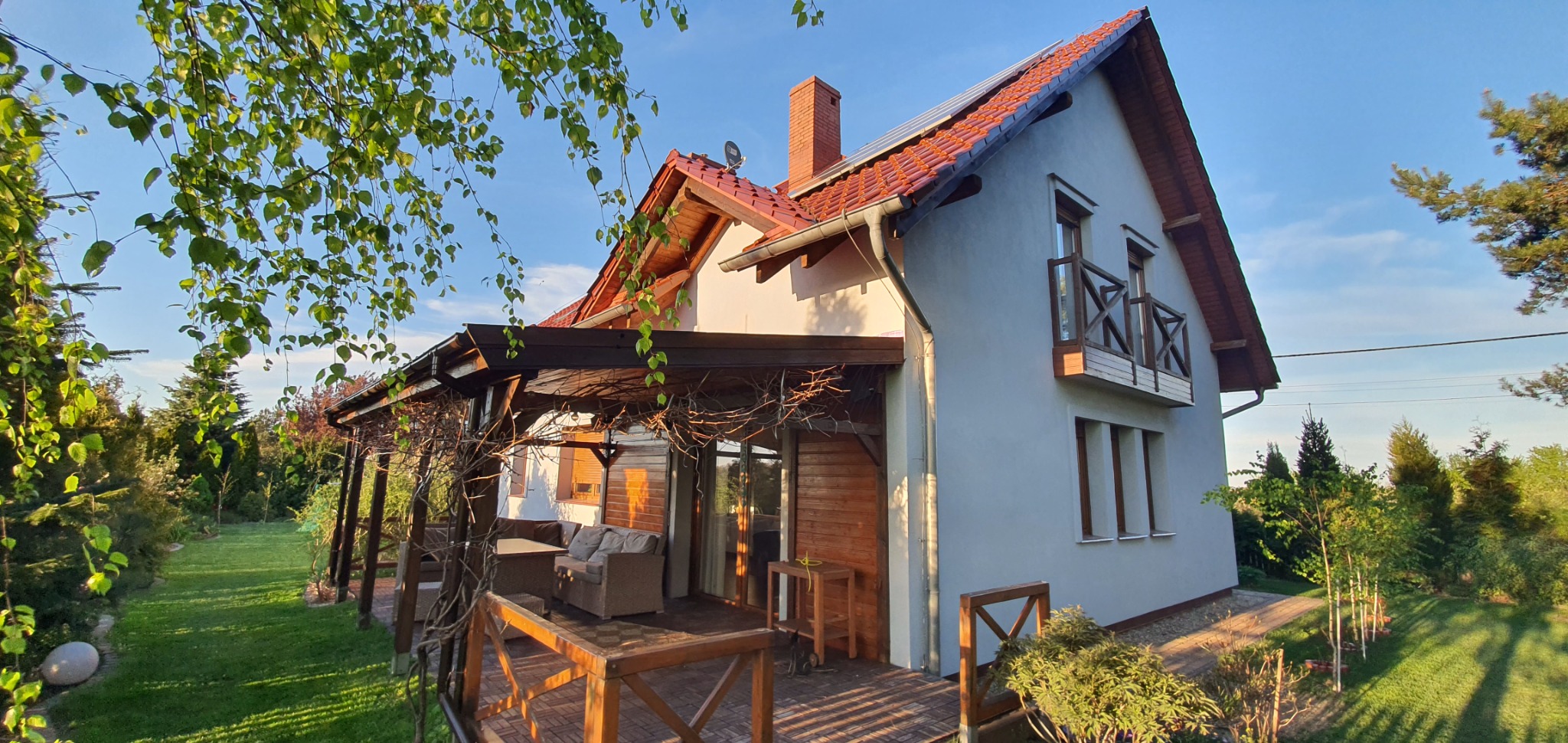 Jasnoszary dom jednorodzinny z czerwoną dachówką, drewnianą werandą i balkonem, otoczony zielonym ogrodem w słoneczny dzień.