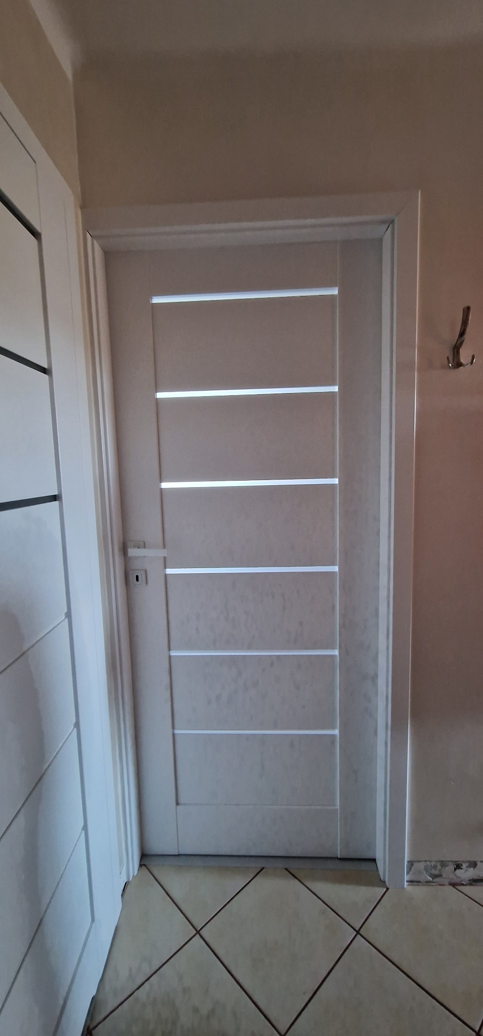 Zamknięte, białe drzwi wewnętrzne z poziomymi przeszkleniami, widoczne z klamką i ościeżnicą, na tle płytek podłogowych i fragmentu ściany z wieszakiem.