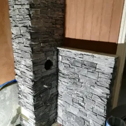 Wykończenie narożnika ściany i obudowy kominka dekoracyjnym kamieniem elewacyjnym w odcieniach szarości, widoczne pozostałości po montażu i taśma malarska.