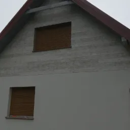Fragment elewacji domu z dwoma oknami z brązowymi roletami zewnętrznymi, g&oacute;rna część elewacji imituje drewno, dolna pomalowana na szaro, widoczny fragment dachu z brązową blachodach&oacute;wką.
