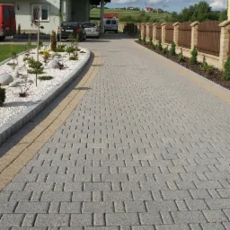 Szeroki podjazd wyłożony szarą kostką z dekoracyjnym obramowaniem z jasnych kostek, obok rabata z białym żwirem i niskimi iglakami, w tle drewniane ogrodzenie i dom.