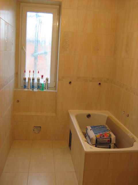 Remont łazienki w trakcie: beżowe płytki, wanna z workami kleju, tuby silikonu na parapecie, otwór w ścianie.