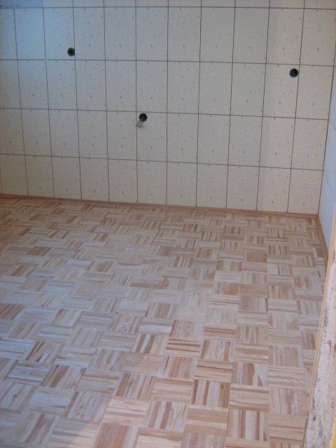 Ułożona parkietowa posadzka w jasnym odcieniu drewna, kremowe płytki ścienne z otworami na instalacje elektryczne i hydrauliczne, widok z góry.