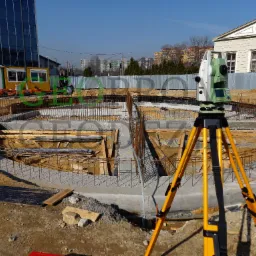 Tachimetr na statywie ustawiony na placu budowy, widoczne zbrojenie fundamentów w kształcie koła, barakowóz i budynki w tle.