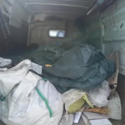 Wnętrze furgonetki wypełnione workami i kartonami z odpadami budowlanymi, widoczne narzędzia na ścianie pojazdu.