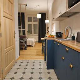 Wnętrze mieszkania po remoncie: widok z przedpokoju na pok&oacute;j dzienny z aneksem kuchennym, charakterystyczne płytki podłogowe w geometryczny wz&oacute;r, drewniana podłoga, niebieskie szafki kuchenne...