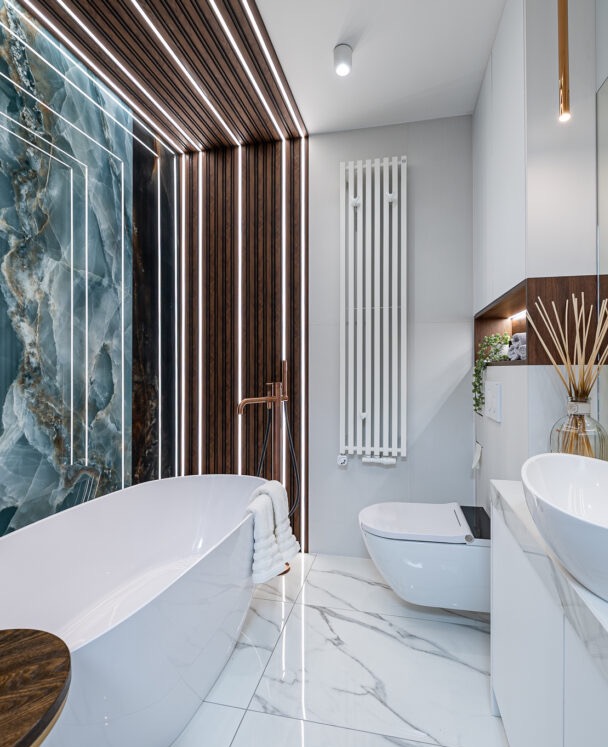 Luksusowa łazienka z wolnostojącą wanną, marmurową podłogą i ścianą z onyksu podświetloną ledami. Nowoczesny design z elementami drewna i armaturą w kolorze różowego złota.