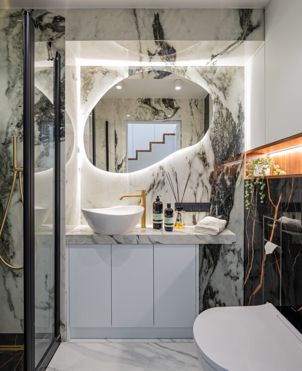 Nowoczesna łazienka z marmurowymi ścianami, owalnym lustrem z podświetleniem LED, białą umywalką nablatową i złotą armaturą, tworząca luksusowy klimat.