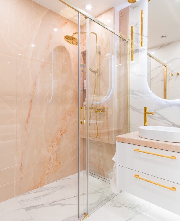 Elegancka łazienka z kabiną prysznicową w odcieniach różu i złota, marmurowe płytki, biała szafka z umywalką i złotymi uchwytami, oświetlenie punktowe.