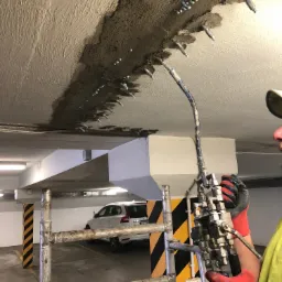 Pracownik w rękawicach aplikuje preparat do naprawy pęknięć na suficie parkingu podziemnego, widoczne kotwy iniekcyjne i ślady wilgoci.