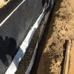 Czarno-biała folia kubełkowa na fundamencie, zabezpieczona geowłókniną i drenażem z kamieni, widoczny fragment pracownika w kombinezonie.