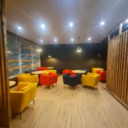 Jasne wnętrze kawiarni lub biura z kolorowymi fotelami (żółte, czerwone, granatowe) i okrągłymi stolikami na jasnej drewnianej podłodze. Widoczne wiszące lampy i okno zabezpieczone folią ochronną.