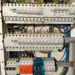 Rozdzielnica elektryczna z zainstalowanymi bezpiecznikami EATON, ogranicznikami przepięć SIMTEC w kolorach pomarańczowym i czerwonym oraz modułami ETI w obudowach turkusowych, widoczne okablowanie...