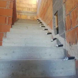 Surowe betonowe schody w trakcie budowy z pomarańczowymi cegłami i czarnymi rurami instalacyjnymi wystającymi ze ścian.