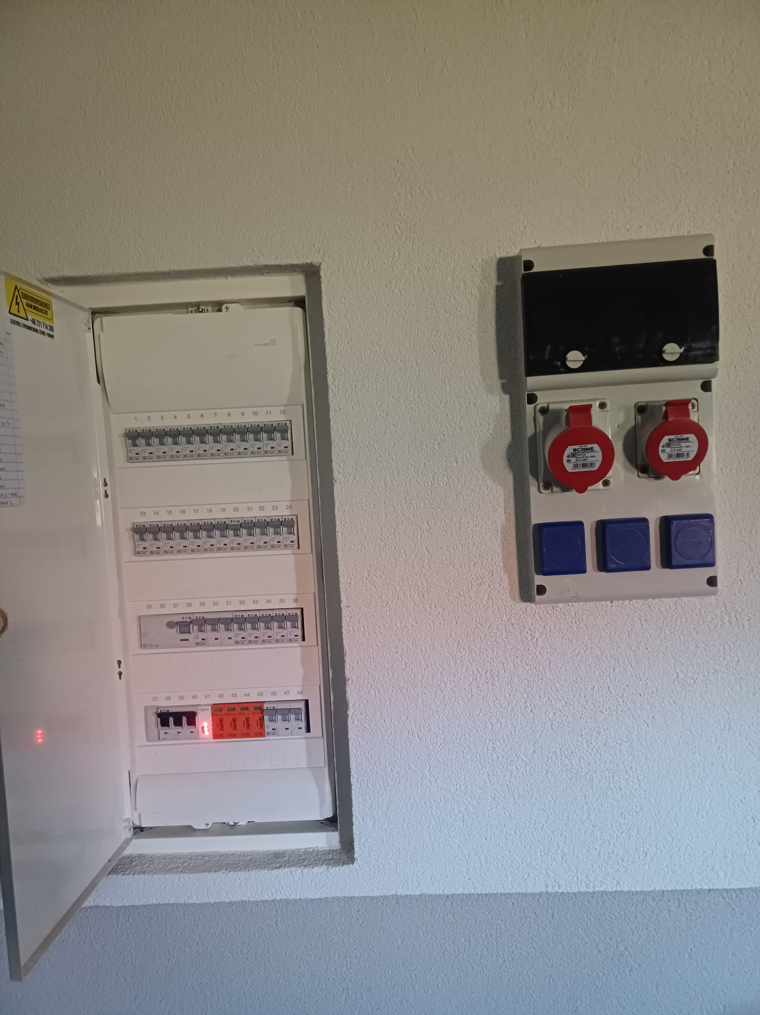 Otwarta skrzynka bezpiecznikowa z widocznymi bezpiecznikami i dwoma włączonymi obwodami, obok puszka z czerwonymi gniazdami siłowymi i niebieskimi gniazdami standardowymi, zamontowane na jasnej...