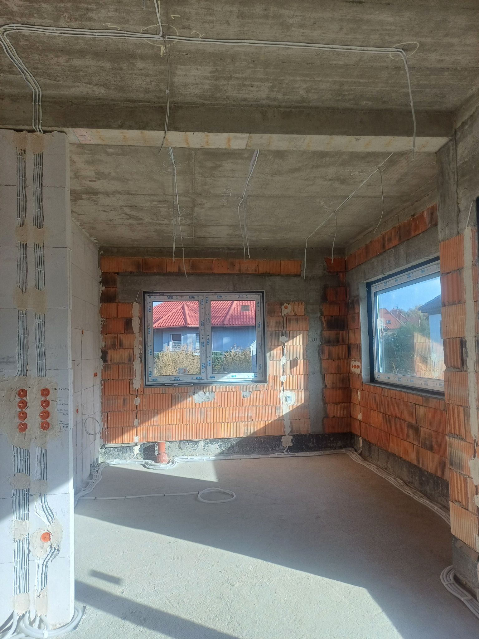 Wnętrze surowego budynku w trakcie prac elektrycznych: widoczne kable prowadzone po suficie i ścianach, puszki instalacyjne w ścianie z gazobetonu, okna z widokiem na dom z czerwoną dachówką.