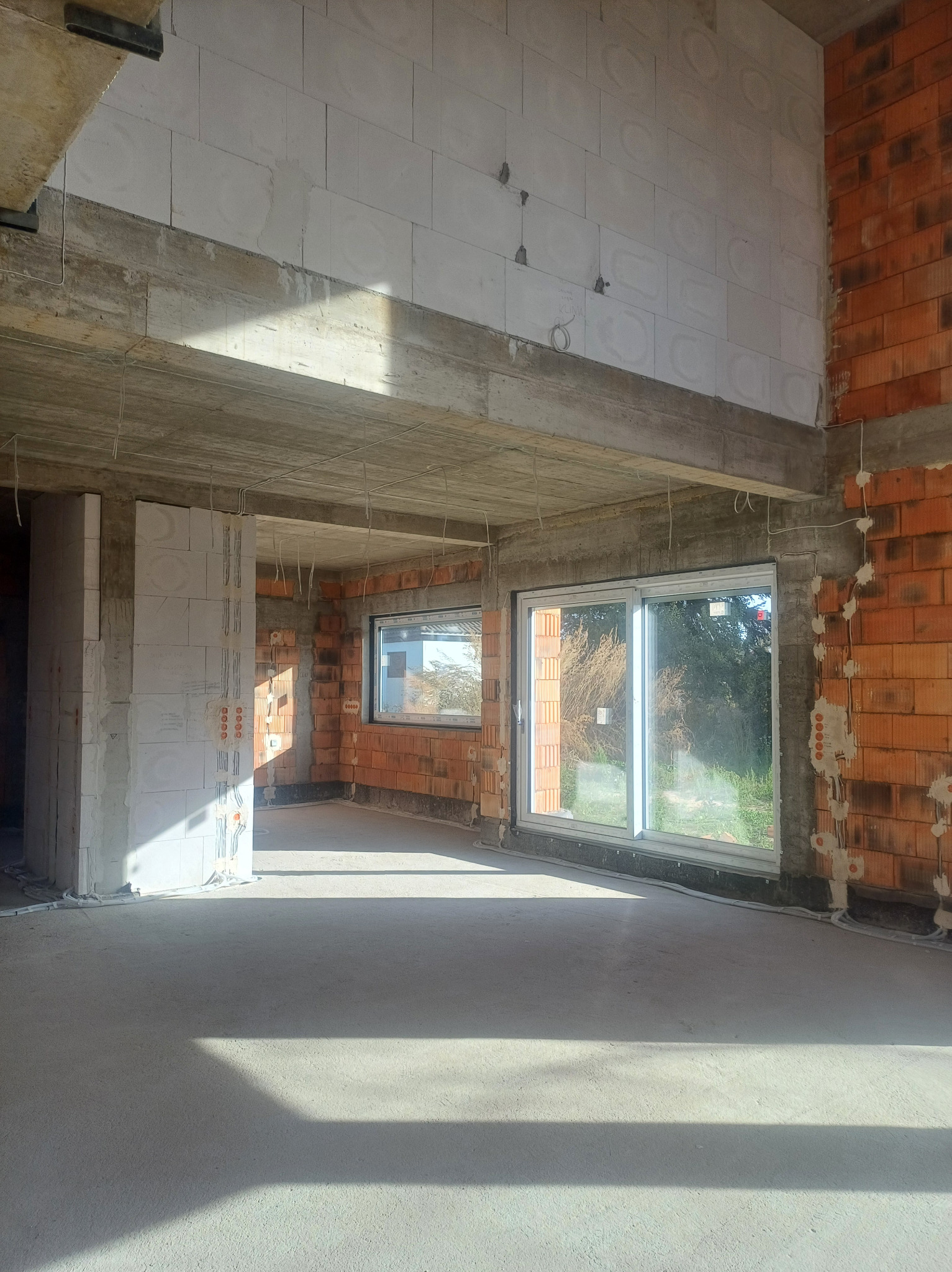 Surowy stan wnętrza domu w trakcie budowy, widoczne ściany z czerwonej cegły i białego betonu komórkowego, betonowy strop, okna, poprowadzone przewody elektryczne w ścianach, naturalne światło...