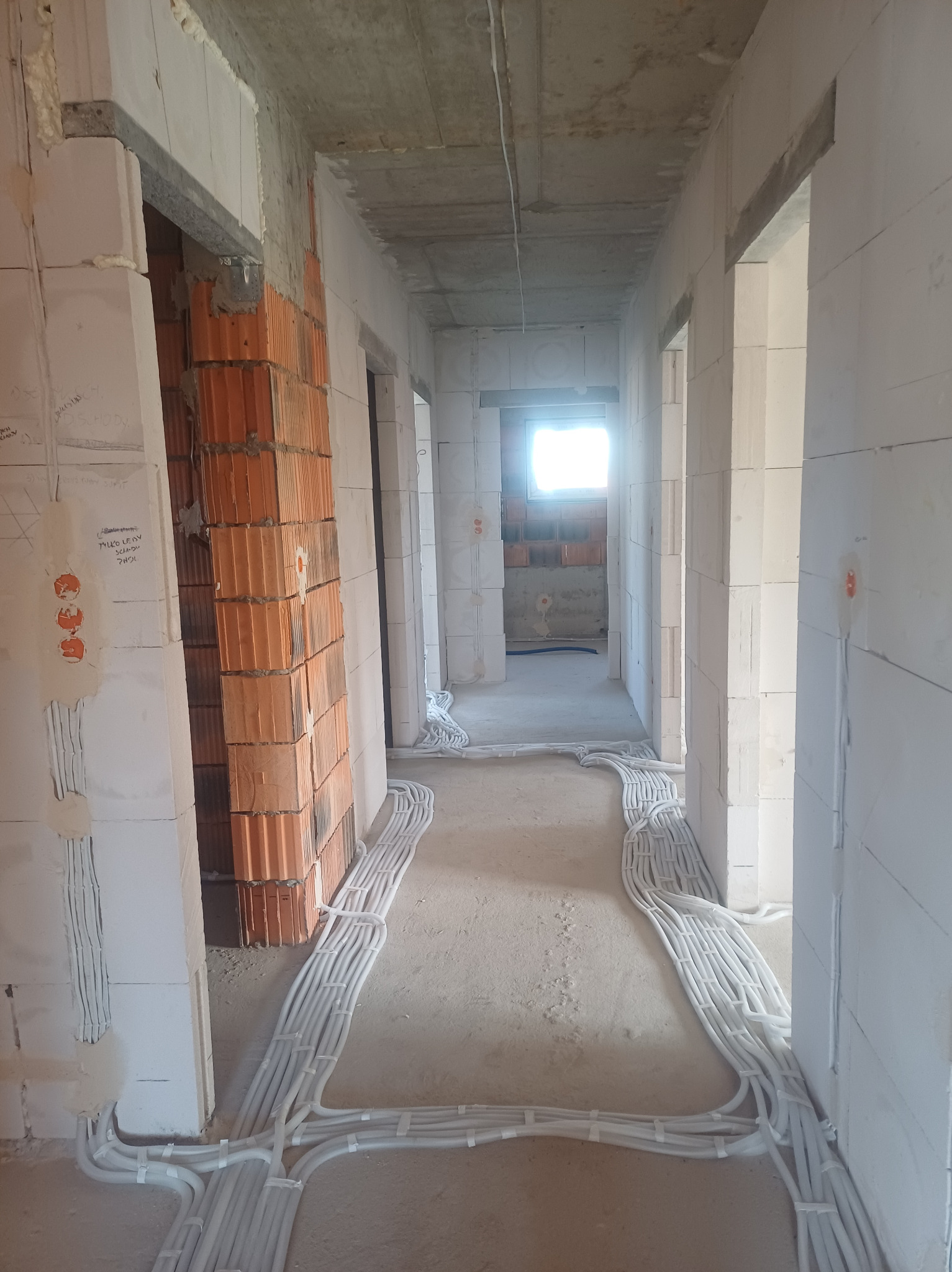 Wnętrze w trakcie budowy, widoczne kable elektryczne prowadzone po podłodze w peszlach, ściany z betonu komórkowego i pustaków ceramicznych, otwory drzwiowe bez ościeżnic.