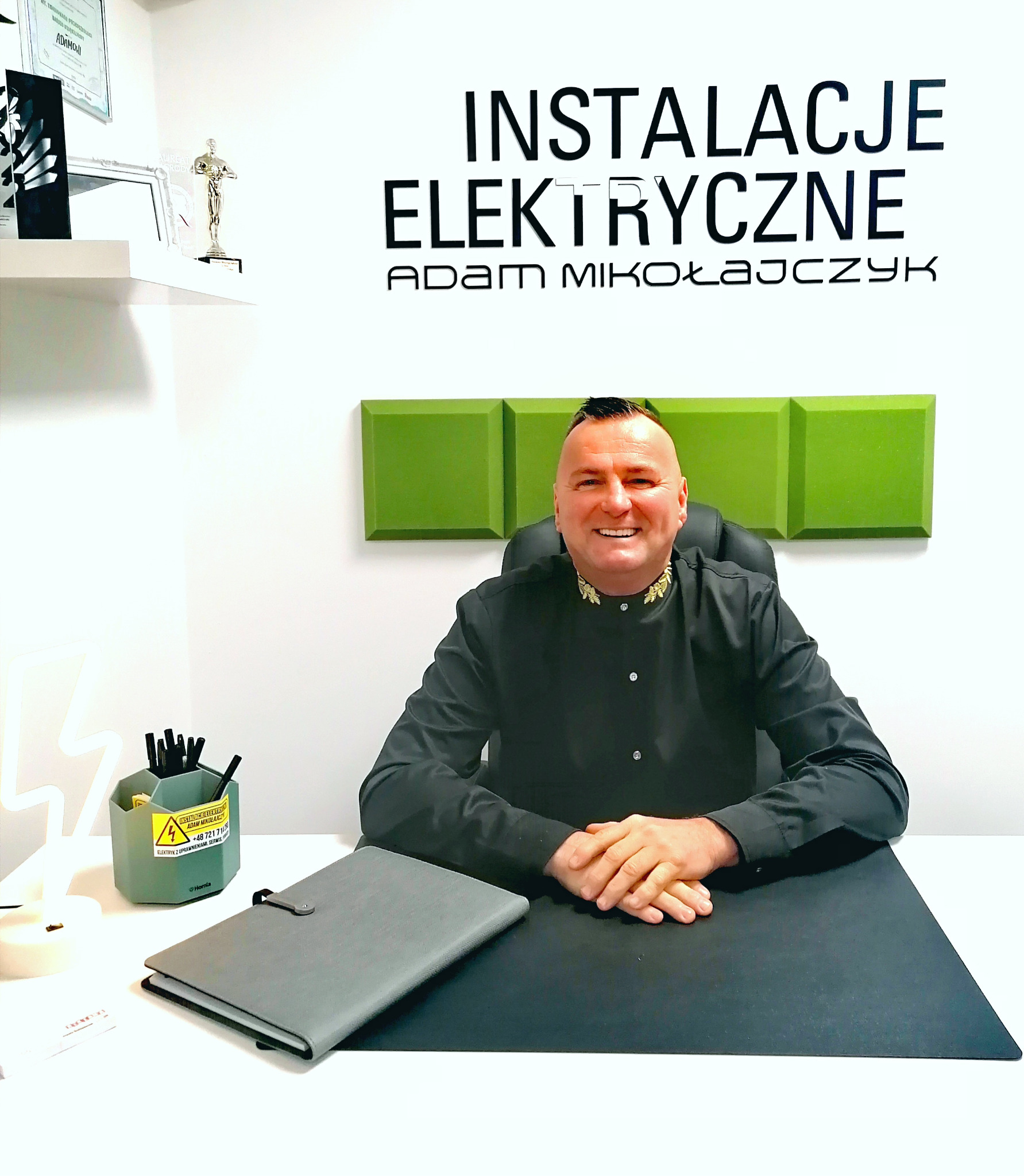 Portret uśmiechniętego mężczyzny w czarnej koszuli siedzącego za biurkiem z napisem 'Instalacje Elektryczne Adam Mikołajczyk' na ścianie w tle. Na biurku organizer z długopisami i notatnik.