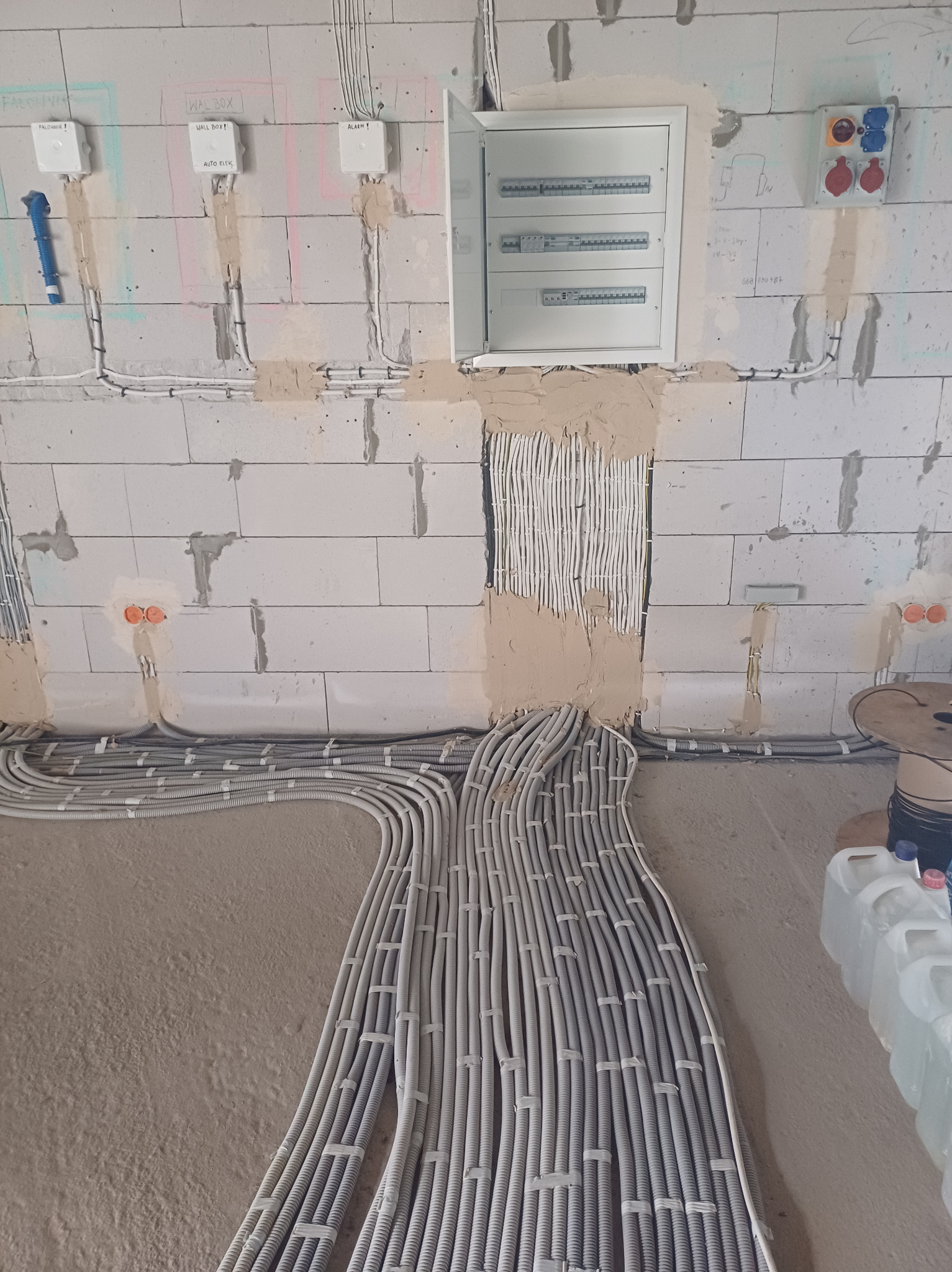 Rozprowadzenie przewodów elektrycznych w peszlach, w trakcie prac instalacyjnych w surowym wnętrzu budynku z widoczną rozdzielnicą i puszkami elektrycznymi w ścianie z betonu komórkowego.