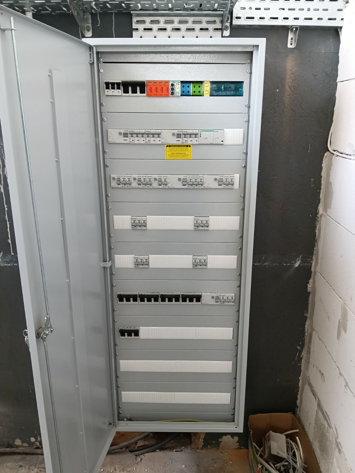Otwarta szafa rozdzielcza z widocznymi zabezpieczeniami i modułami, zamontowana na ścianie w surowym stanie, z widocznymi przewodami u dołu.