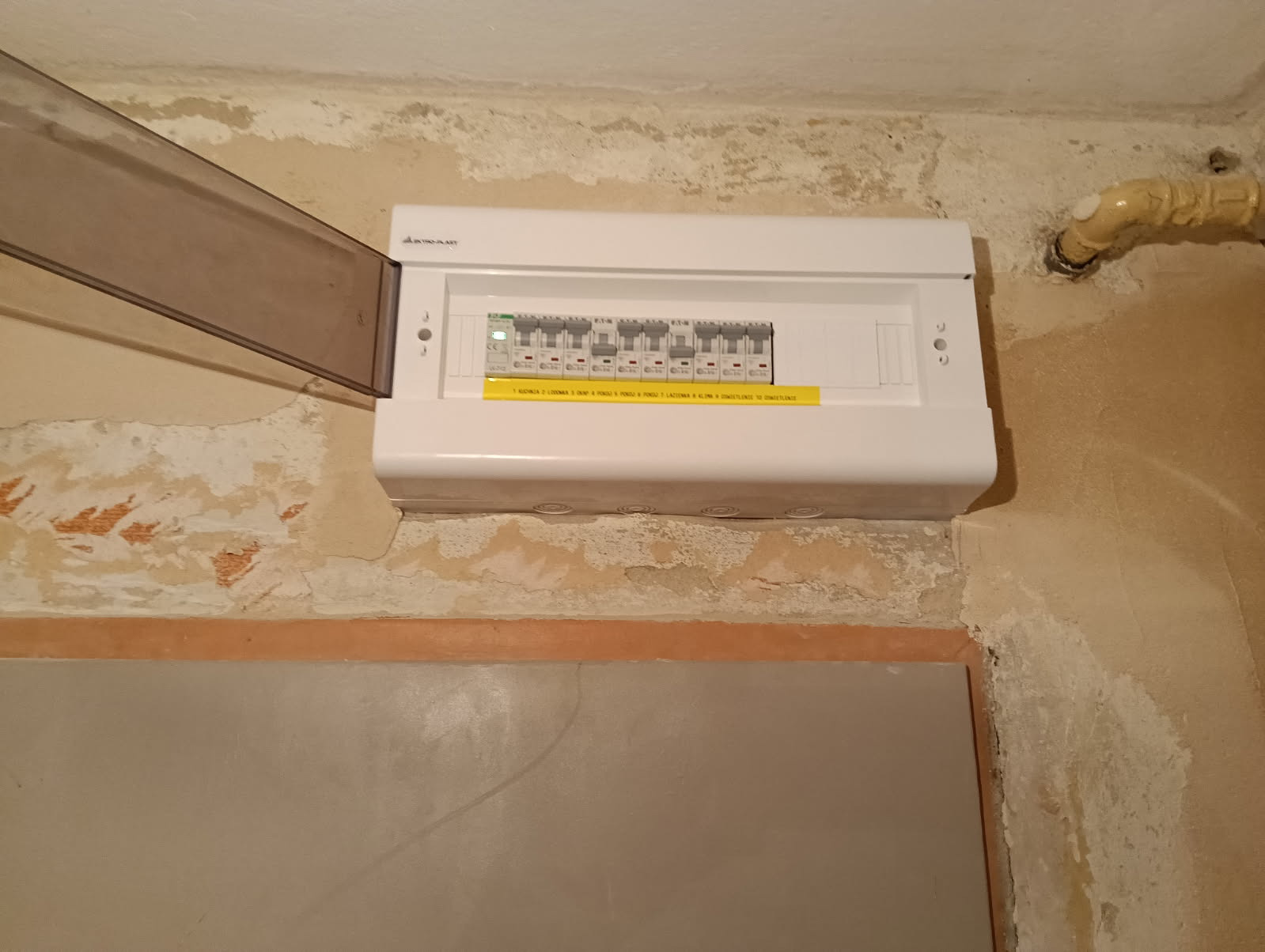 Biała skrzynka rozdzielcza z bezpiecznikami na ścianie z odchodzącą farbą, z otwartą, przezroczystą klapką. Widoczny fragment rury.