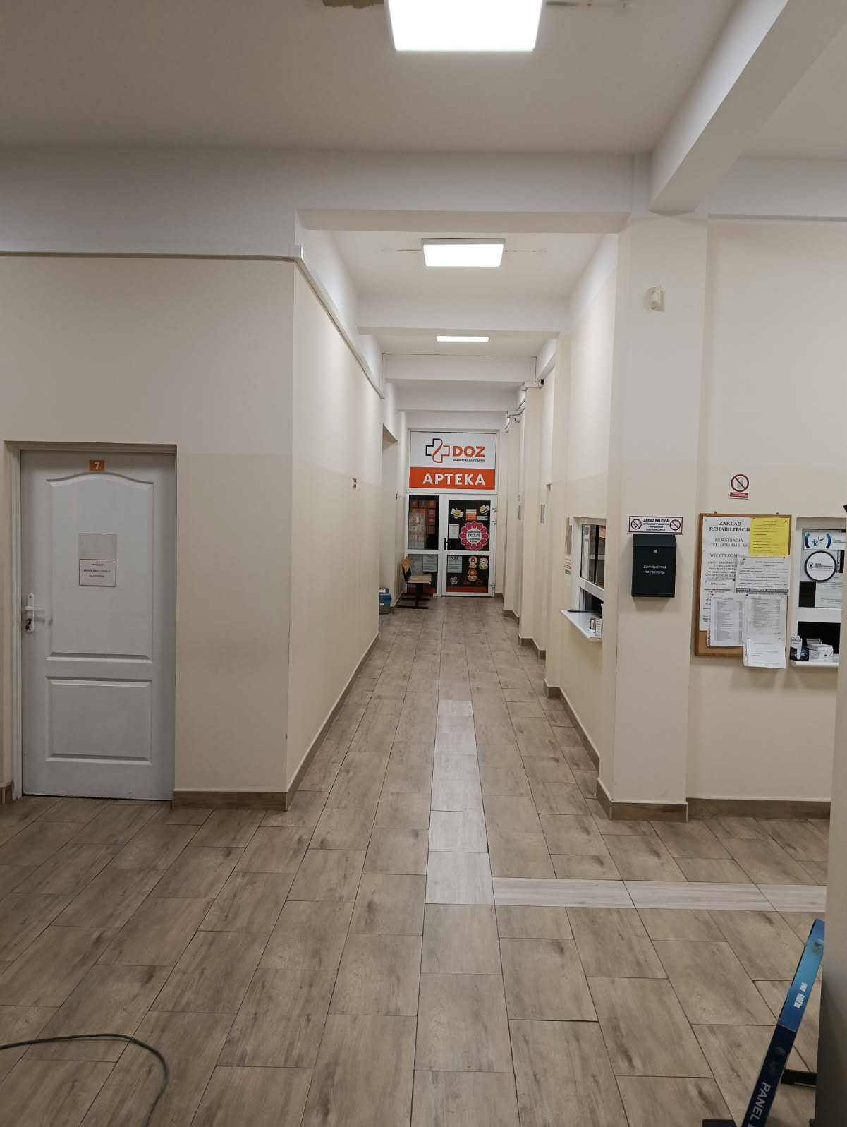 Długi, pusty korytarz z beżowymi ścianami i brązowymi płytkami podłogowymi prowadzący do apteki DOZ. Widoczne tablice informacyjne i drzwi po bokach.