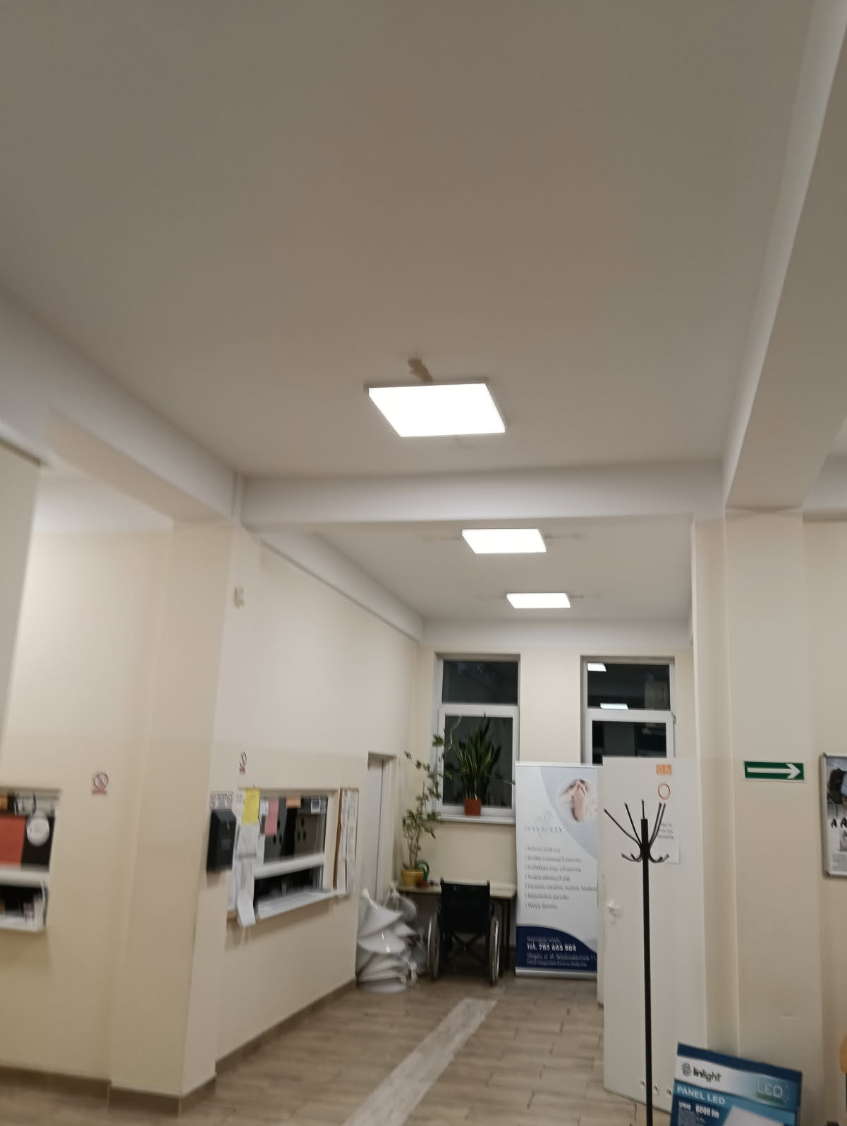 Kwadratowe panele LED w podwieszanym suficie korytarza, oświetlające wnętrze z roślinami, wózkiem inwalidzkim i punktem obsługi klienta.