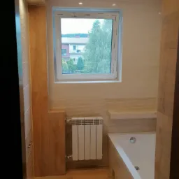 Jasna łazienka w Mucharzu z beżowymi płytkami imitującymi drewno, biała wanna i grzejnik pod oknem z widokiem na zieleń.