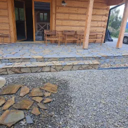 Wykończony taras z kamienia naturalnego przy drewnianym domu, z fragmentami kamienia do układania na żwirowym podłożu, widoczne drewniane krzesła i fragment samochodu.