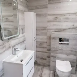 Nowoczesna łazienka w odcieniach szarości i bieli z umywalką nablatową, podświetlanym lustrem LED, wiszącą toaletą i szafką łazienkową, wykończona płytkami imitującymi marmur.